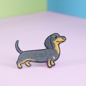 Peut inclure: Patch brodé thermocollant représentant un teckel noir et fauve, debout sur une surface rose.