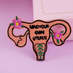 May include: A pink embroidered patch with a uterus shape and the text "Mind your own uterus" in black. The uterus is decorated with two pink flowers and green leaves.