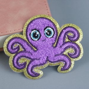 Op de afbeelding: Een paarse octopus patch met een gouden glitterrand. De octopus heeft grote, expressieve ogen met blauwe en witte details. De patch is geborduurd en heeft een getextureerde uitstraling, perfect om toe te voegen aan kleding of accessoires.
