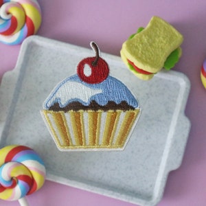 Puede incluir: Un cupcake azul y blanco con una cereza roja en la parte superior, bordado en una bandeja blanca. El cupcake está rodeado de piruletas de colores, una hamburguesa y un sándwich.
