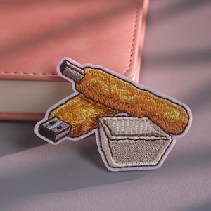 Puede incluir: Un parche bordado que muestra un alimento frito de color dorado, posiblemente un churro o un palito de pan, colocado en diagonal sobre un pequeño recipiente cuadrado de color claro. El parche tiene un borde blanco y costuras detalladas.