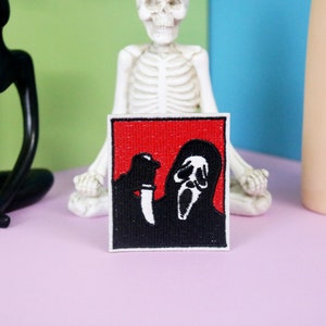 May include: A black and white embroidered patch of a masked figure holding a knife. The figure is wearing a white mask with a scream face. The background is red.