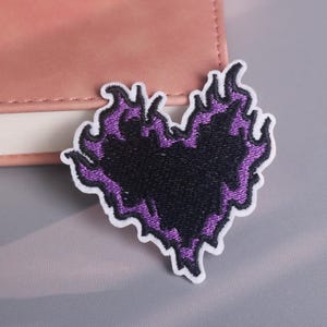 Puede incluir: Parche bordado en forma de corazón con centro negro y llamas moradas. El parche tiene un borde blanco y está colocado sobre una superficie gris claro junto a un cuaderno rosa.