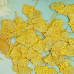 May include: A collection of yellow ginkgo leaf embellishments, intricately crafted with a textured, beaded appearance. The leaves are arranged on a light blue surface, with white daisies in the upper left corner. These decorative elements are ideal for craft projects.