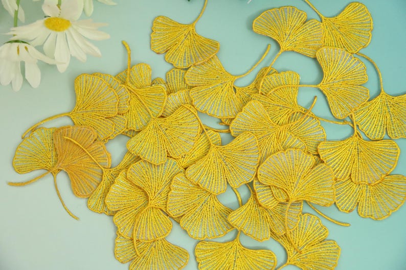 May include: A collection of yellow ginkgo leaf embellishments, intricately crafted with a textured, beaded appearance. The leaves are arranged on a light blue surface, with white daisies in the upper left corner. These decorative elements are ideal for craft projects.