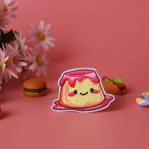 May include: A pink and yellow embroidered patch of a cartoon pudding with a happy face. The pudding is surrounded by other small toy foods, including a hamburger, a hot dog, and a lollipop.