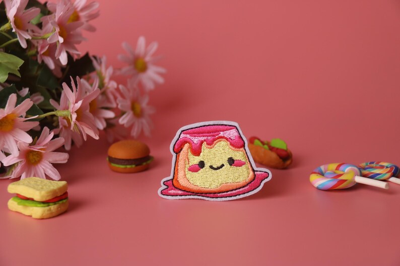 May include: A pink and yellow embroidered patch of a cartoon pudding with a happy face. The pudding is surrounded by other small toy foods, including a hamburger, a hot dog, and a lollipop.