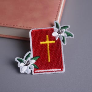 May include: Embroidered patch featuring a red book with a yellow cross, accented by white and green floral details. The patch is rectangular with a white border. The design is set against a neutral gray surface.
