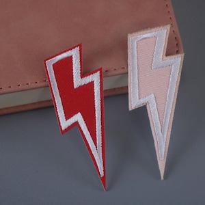 Op de afbeelding: Twee strijkbare patches in de vorm van een bliksem. Eén patch is rood met witte rand, de andere is roze met witte rand.