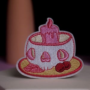 Può includere: Toppa ricamata raffigurante una tazza a forma di teschio con una candela rosa e due ciliegie rosse. La toppa ha una combinazione di colori bianco, rosa e oro. Il design è dettagliato con elementi cuciti.