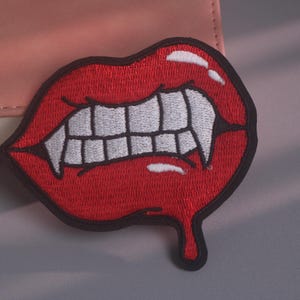 Puede incluir: Un parche bordado que muestra un par de labios de color rojo brillante con colmillos blancos mordiendo el labio inferior. El diseño incluye reflejos blancos en los labios y un efecto de goteo rojo en la parte inferior, todo con un borde negro.