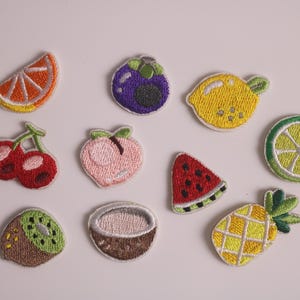 Puede incluir: Parches bordados de frutas, incluyendo una rodaja de naranja, un mangostán morado, un limón amarillo, cerezas rojas, un melocotón rosa, una rodaja de sandía, una rodaja de lima, un kiwi, un coco y una piña. Parches coloridos y detallados.