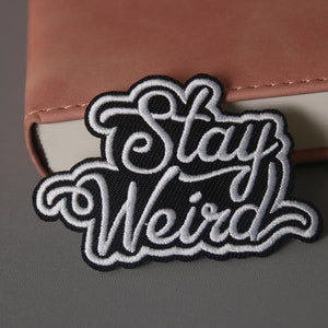 Op de afbeelding: Zwarte geborduurde patch met witte tekst "Stay Weird".