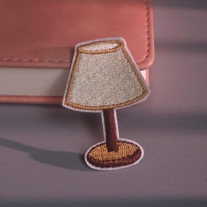 May include: An embroidered patch shaped like a table lamp with a beige lampshade, a dark brown stem, and a golden-brown circular base. The patch features a white border outline and is positioned against a pink rectangular object.