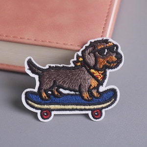 May include: Embroidered patch featuring a dachshund dog wearing sunglasses and a bandana, riding a blue skateboard with red wheels. The patch has a white border and is set against a gray surface.