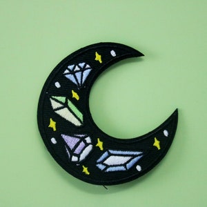 Puede incluir: Parche de luna creciente negro con cristales blancos, azules, verdes y morados bordados y estrellas amarillas.