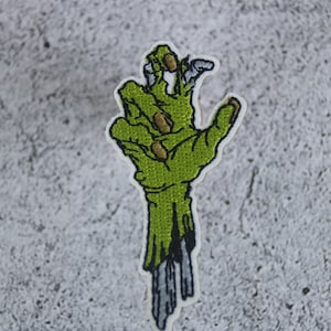 May include: Green zombie hand with white stitching, reaching out with fingers outstretched. The hand is partially decayed with grey stitching representing the flesh falling off.