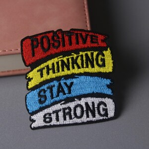Op de afbeelding: Een kleurrijk geborduurd patch met de woorden "POSITIVE THINKING STAY STRONG" in zwarte letters op een rode, gele, blauwe en witte achtergrond.