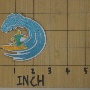 Może przedstawiać: Naszywka haftowana do prasowania przedstawiająca surfera jadącego na fali. Fala jest niebieska i biała, a surfer ma na sobie zieloną koszulę i niebieskie spodenki. Deska surfingowa jest ż&oacute;łta.