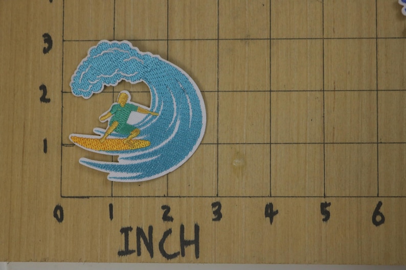 Może przedstawiać: Naszywka haftowana do prasowania przedstawiająca surfera jadącego na fali. Fala jest niebieska i biała, a surfer ma na sobie zieloną koszulę i niebieskie spodenki. Deska surfingowa jest ż&oacute;łta.