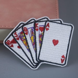 Könnte beinhalten: Ein weißer gestickter Patch mit fünf Spielkarten, darunter das Ass Pik, der König Herz, die Dame Herz, der Bube Herz und die Zehn Herz. Die Karten sind fächerförmig angeordnet, wobei das Ass Pik oben liegt.