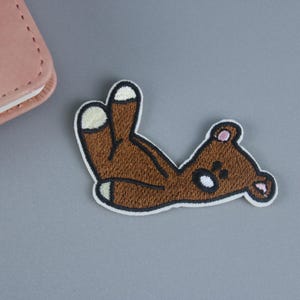 May include: Brown embroidered iron-on patch of a teddy bear lying on its back with its paws in the air.