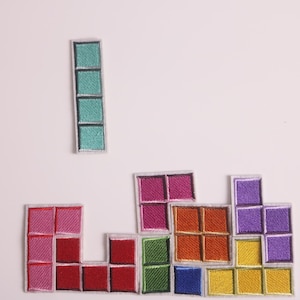 May include: Embroidered patches in the style of Tetris game pieces. The patches are in various colors, including teal, pink, red, green, orange, blue, purple, and yellow. The patches are arranged to resemble a Tetris game.