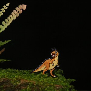 May include: A small, orange and purple embroidered dinosaur patch with a toothy grin, sitting on a bed of green moss.