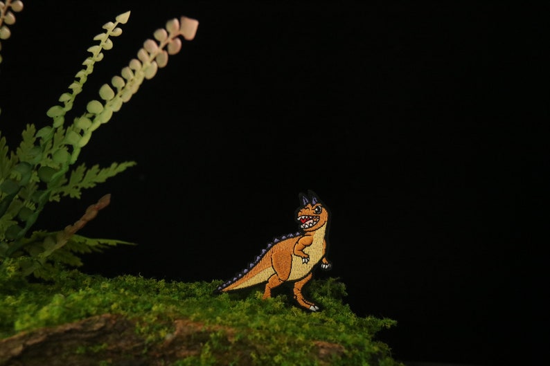 May include: A small, orange and purple embroidered dinosaur patch with a toothy grin, sitting on a bed of green moss.
