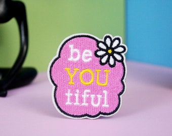 Könnte beinhalten: Rosa gestickter Patch mit einer weißen Gänseblümchen und dem Text "be you tiful" in weißen und gelben Buchstaben.