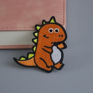 May include: Embroidered patch featuring a cartoon dinosaur in orange, yellow, and black. The dinosaur has a white belly and is standing. The patch is likely for clothing or accessories.