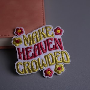 May include: Embroidered patch with the words "MAKE HEAVEN CROWDED" in yellow and red, surrounded by small flower accents. The patch is on a white background. A pink notebook is in the background.