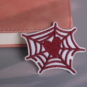 May include: Embroidered patch featuring a spider web design with a heart in the center. The patch is primarily white with burgundy-colored details. The patch is placed on a pink leather-bound notebook.