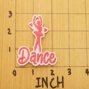 Op de afbeelding: Geborduurde patch met een roze ballerina en het woord "Dance". De patch bevindt zich op een houten oppervlak met inch-maten. De patch is ongeveer 5,08 cm lang.