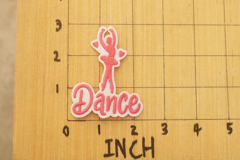 Op de afbeelding: Geborduurde patch met een roze ballerina en het woord "Dance". De patch bevindt zich op een houten oppervlak met inch-maten. De patch is ongeveer 5,08 cm lang.