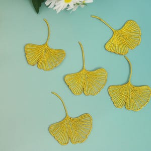 May include: Five gold-colored ginkgo leaf appliques are arranged on a light blue background. The leaves have a detailed, openwork design. White flowers are visible in the upper left corner.