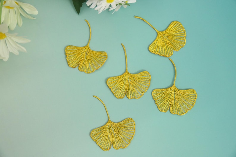 May include: Five gold-colored ginkgo leaf appliques are arranged on a light blue background. The leaves have a detailed, openwork design. White flowers are visible in the upper left corner.