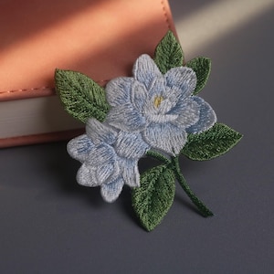 May include: Embroidered floral patch featuring two light blue flowers with yellow centers and green leaves. The patch is detailed with intricate stitching, creating a textured appearance. The patch is likely for decorative purposes, such as adorning clothing or accessories.