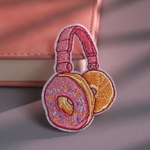 Op de afbeelding: Een geborduurde patch met een koptelefoon waarbij de oorschelpen de vorm hebben van donuts. De ene donut is roze met gekleurde hagelslag en de andere is goudbruin. De hoofdband is roze en verbindt de twee donutvormige oorschelpen met elkaar.