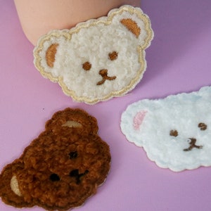 Könnte beinhalten: Drei Plüsch-Bären-Patches in verschiedenen Farben: Weiß, Braun und Hellblau. Die Patches sind mit Gesichtszügen bestickt und haben eine flauschige Textur.