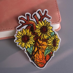 Puede incluir: Parche bordado con un diseño de corazón anatómico estilizado con girasoles. El corazón está representado en tonos naranja y rojo, con girasoles amarillos y hojas verdes. El parche tiene un borde blanco.