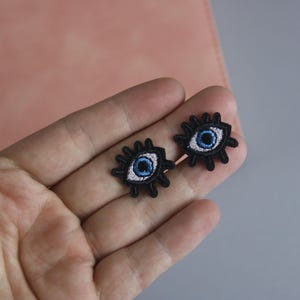 May include: Two black embroidered eye patches with blue and white irises. The patches are on a hand.