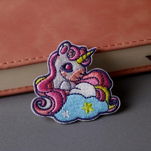 May include: An embroidered unicorn patch in pink and white with a yellow horn and a blue cloud with yellow stars. The patch is on a grey surface.