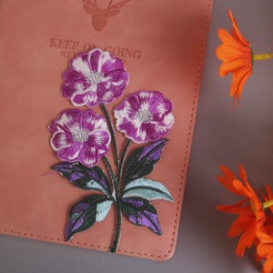 Pode incluir: Um caderno cor-de-rosa com um desenho floral bordado com flores roxas e brancas e folhas verdes e roxas. O caderno tem as palavras "KEEP ON GOING" impressas. Flores laranja no fundo.