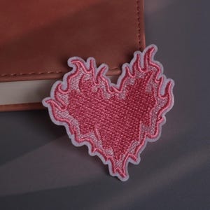 Pode incluir: Patch bordado rosa em forma de coração com chamas nas bordas e borda branca. O patch está em uma superfície cinza ao lado de um caderno marrom.
