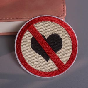 May include: Embroidered patch with a red border and a red diagonal line through a black heart symbol. The patch has a beige background and a white border. The patch is round and is placed on a gray surface.