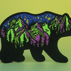 May include: Black embroidered patch of a bear silhouette with a mountain range and starry night sky inside. The mountains are green, purple, and blue with gold stars and a crescent moon.