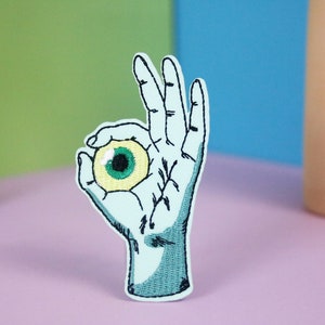 May include: A blue embroidered patch of a hand making the OK sign, holding a green and yellow eyeball.