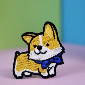 Op de afbeelding: Gestikte patch van een cartoon corgi met een blauwe strik. De corgi is geel met witte markeringen en zwarte ogen.