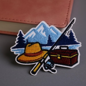 Könnte beinhalten: Gestickter Aufnäher mit Angelmotiv. Das Design umfasst einen gelben Hut, eine Angelrute, eine Angelkiste und eine Bergkette mit Kiefern. Der Aufnäher hat einen weißen Rand und ist vor grauem Hintergrund platziert.